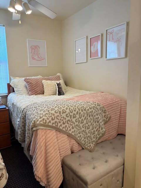 a room decorate with white and pink accents. There are light pink graphics on the wall in white frames. White bedding with a pink and leopard throw blanket and a grey ottoman at the foot of the bed.